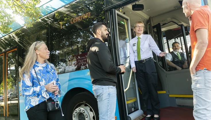 Bussförare står i öppningen till en buss och hälsar passagerare välkomna ombord.