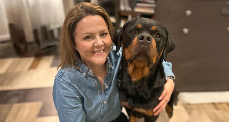 Person i jeansskjorta sitter bredvid en stor svart och brun hund inomhus. Personen håller armen om hunden, som sitter nära och är vänd mot kameran.
