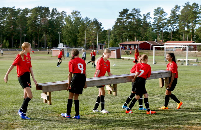 Några spelare ur ett flicklag i fotboll som bär på en bänk