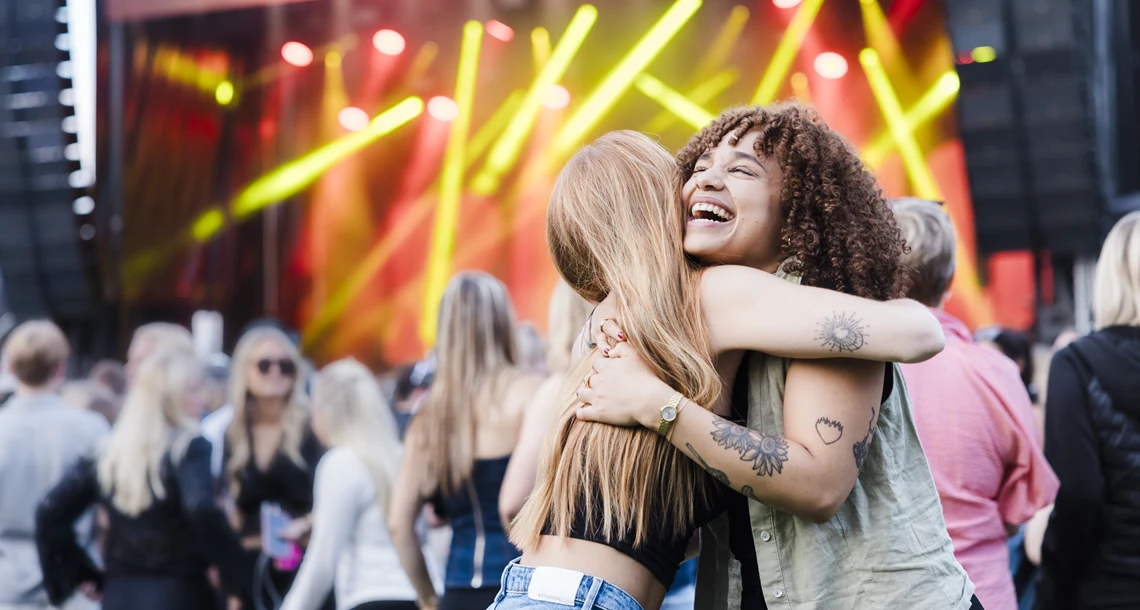 Två tjejer på konsert med pubilk och scen i bakgrunden.