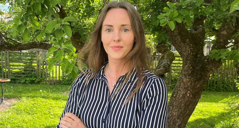 Person med axellångt hår står utomhus framför ett träd med gröna blad, armarna korsade. Personen bär en mörk, randig skjorta och en tunn halskedja. I bakgrunden syns en gräsmatta, ett trästaket och lummig trädgård i dagsljus.