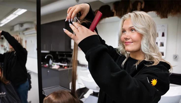 En tjej klipper en annan tjej framför en spegel i salongsmiljö.