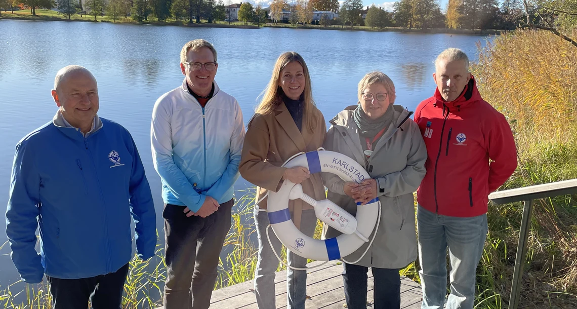 Utomhus. Porträtt. Fem personer står på brygga. I bakgrunden vatten och sol.