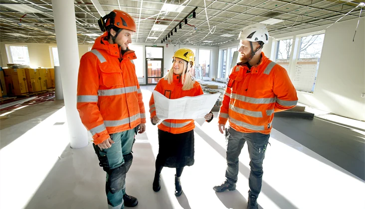 Bild på tre personer med arbetskläder i byggmiljö.