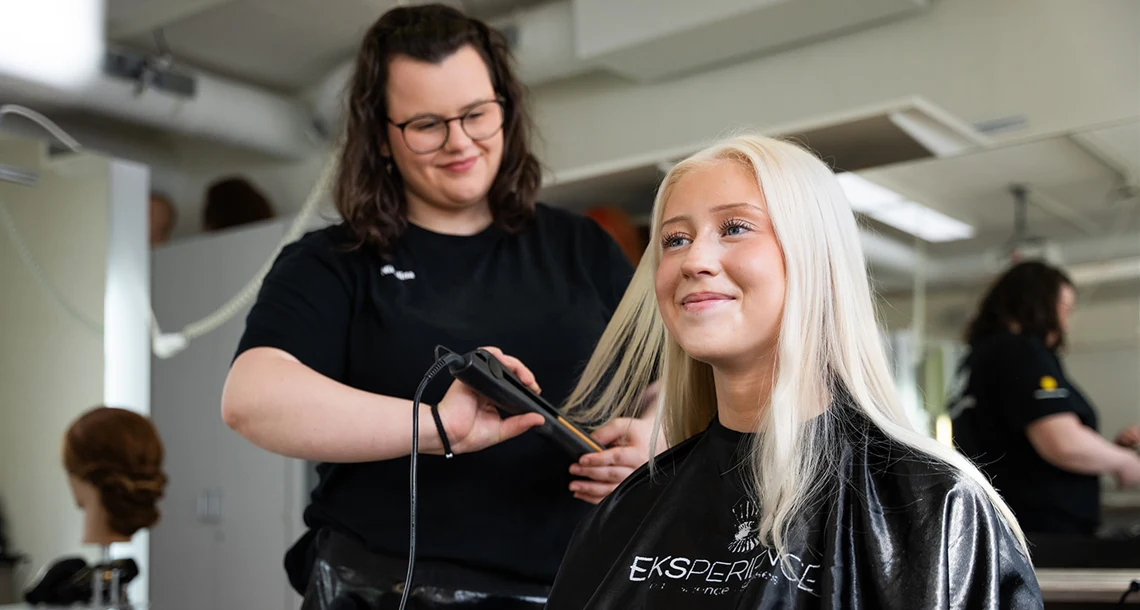 En tjej står upp och fixar håret med en plattång på en annan tjej som sitter ner. De ser glada ut.