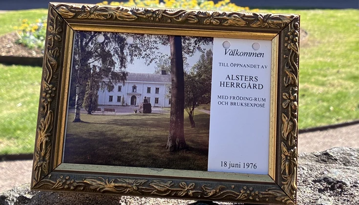 Inramat vykort med inbjudan till öppnandet av Alsters herrgård 1976. Vit herrgårdsbyggnad i bakgrunden. 