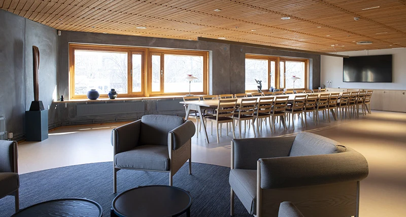 I förgrunden står ett par fåtöljer och i bakgrunde ett långt mötesbord med stolar. Bakom bordet är en lång vägg med fönster.