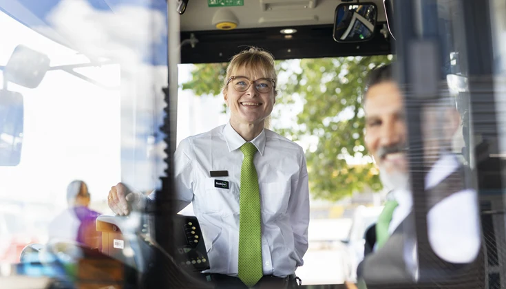 Två bussförare tittar ut genom ett fönster i en buss och ler.