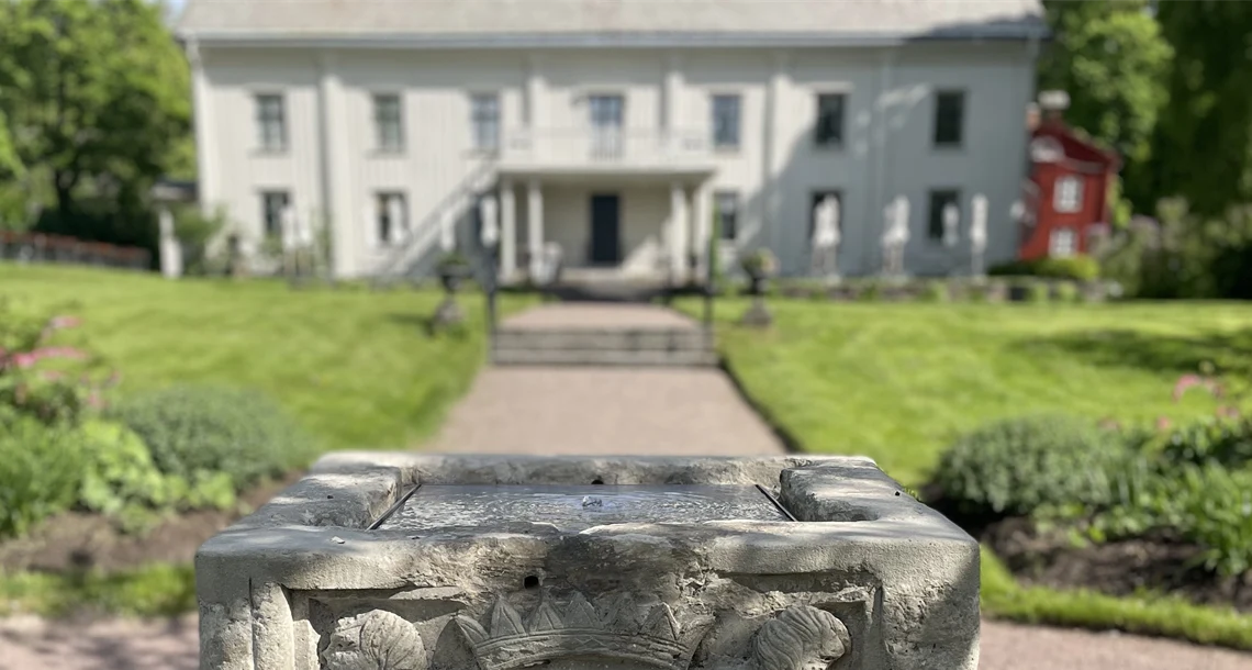 Stenbrunn i förgrunden med park och vit byggnad i bakgrunden