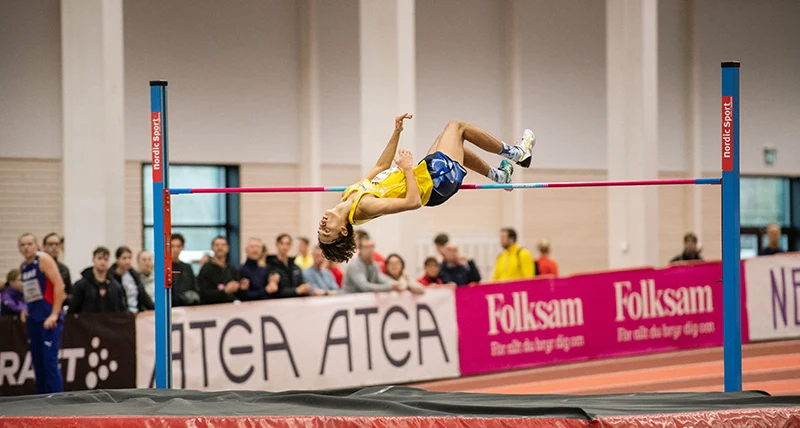 En killa i gula sportkläder hoppar höjdhopp och det står publik i bakgrunden.