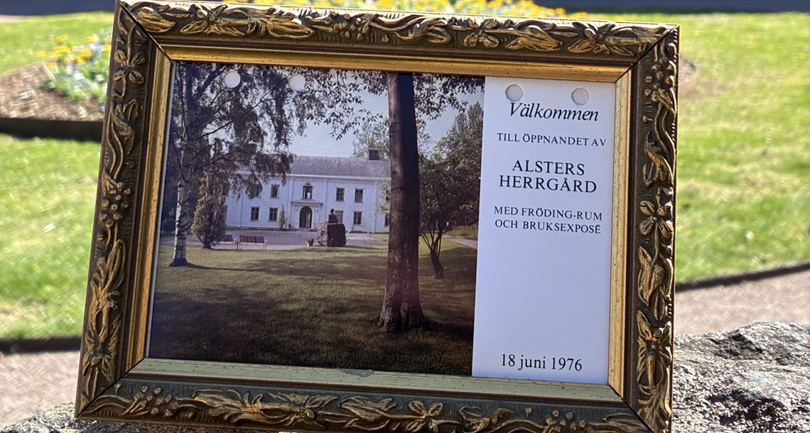 Inramat vykort med inbjudan till öppnandet av Alsters herrgård 1976. Vit herrgårdsbyggnad i bakgrunden. 