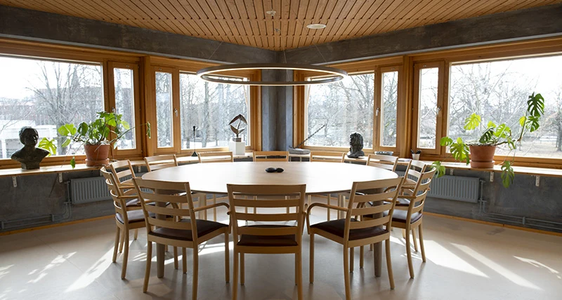 I hörnet av ett rum med fönster åt båda håll står ett runt bord med flera stolar runtom.