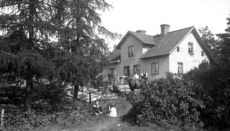 Ett svartvitt foto på en trädgård med flera personer i och en häst. I bakgrunden syns en stenhus.