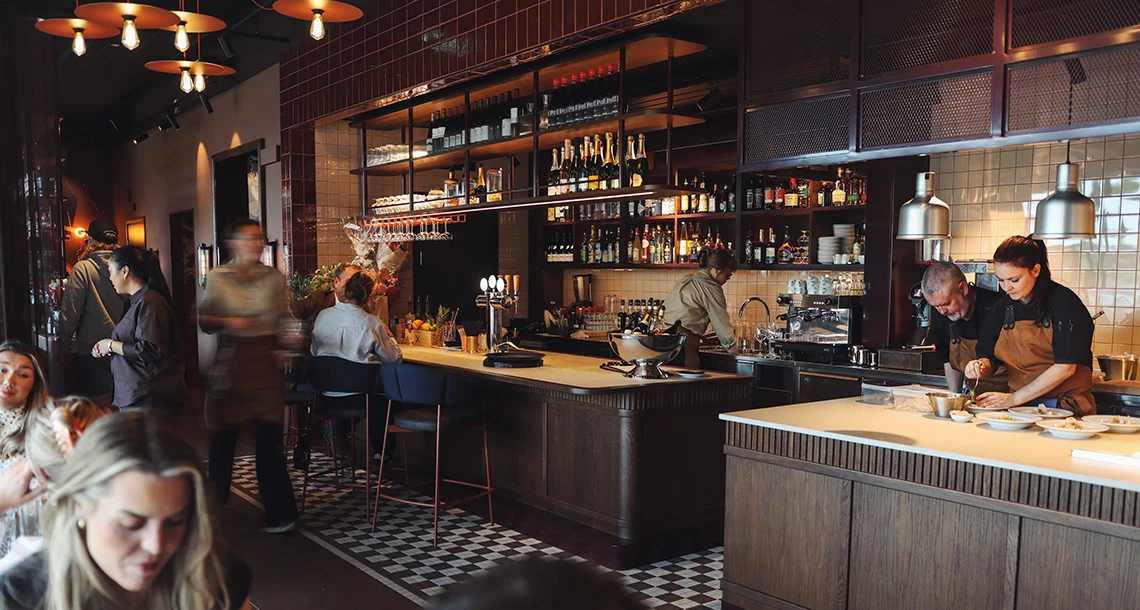 Interiör från en restaurang med diskar i mörkt trä, ätande gäster och ett schackrutigt golv.