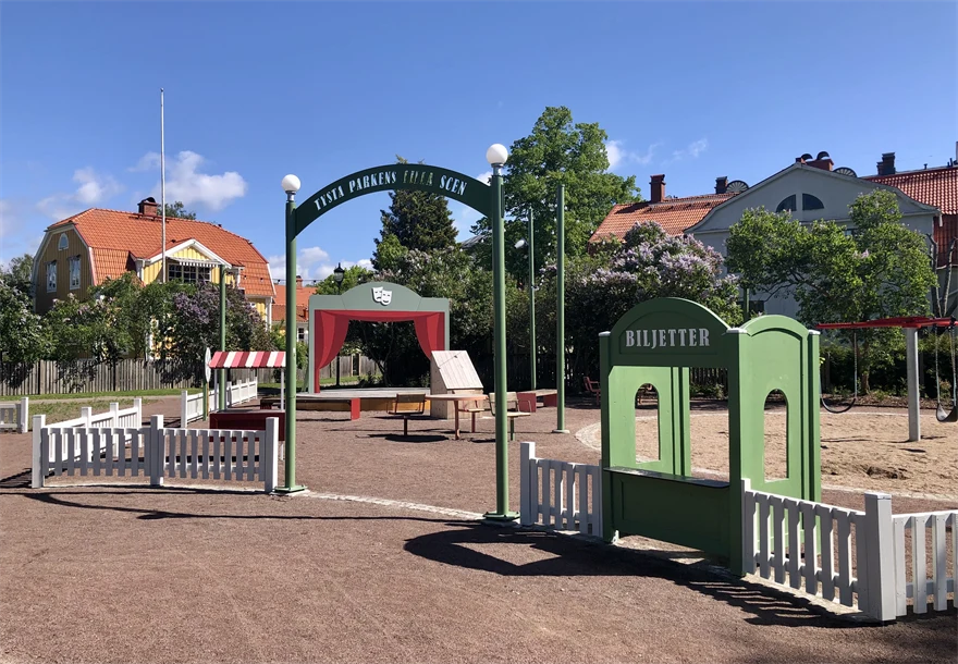 Bakom en vitt staket syns en liten scen med trädraperier och en biljettlucka.