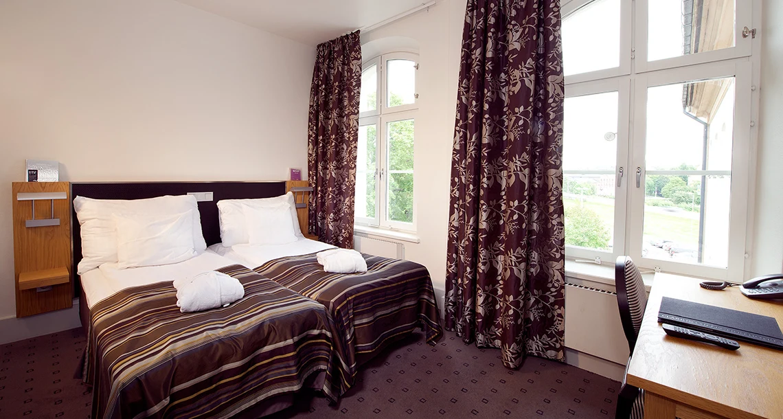 En dubbelsäng med randiga bruna överkast framför två stora välvda fönster