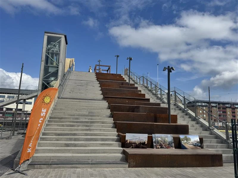 Trappa med breda steg och integrerade sittplatser. Intill en hiss och ett orange informationsflagg med text. Vid trappan visas fotografier och i bakgrunden syns stadsmiljö och blå himmel med moln.