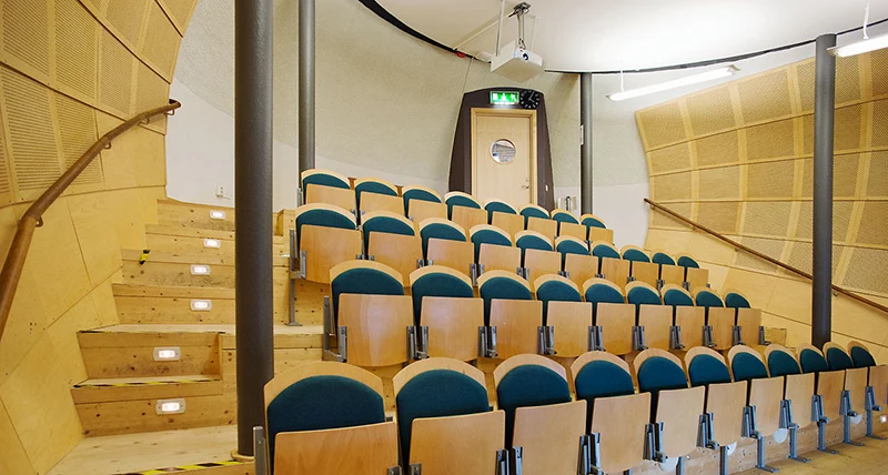 Fyra rader med stolar i en lokal med brant lutning mellan stolsraderna. Väggarna är välvda inåt.